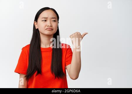 Lugubre fille chinoise fronde des sourcils, points et regarde à droite avec déception, se trouve frustré contre fond blanc Banque D'Images