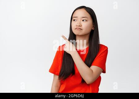 Déçue asiatique jeune fille montre le regret, pointant et regardant à gauche avec le visage de frowning, se tenant bouleversé sur fond blanc Banque D'Images