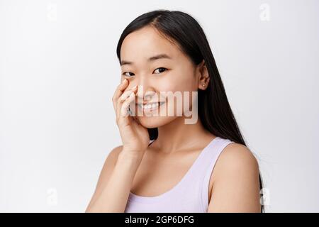 Belle jeune femme asiatique avec une peau saine et éclatante, visage souriant et touchante avec le bout des doigts, debout sur fond blanc Banque D'Images