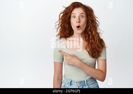 Image de la fille de tête rouge qui souffle et qui semble stupéfait, pointant vers la gauche avec l'expression du visage surprise, fond blanc Banque D'Images