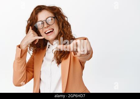 Employés de bureau. Souriante fille rouge en costume pointant le doigt vers l'appareil photo, montrant le geste du téléphone mobile, lui demandant de lui donner un appel, vous recrutant Banque D'Images