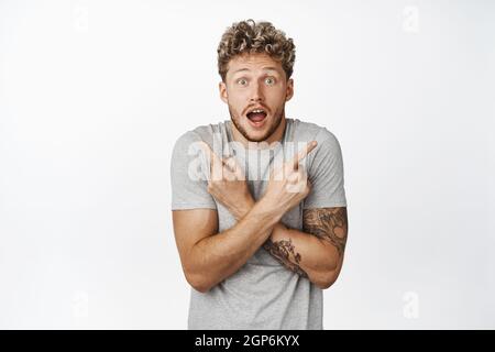 Surpris homme blond pointant les doigts sur le côté, regardant indécise et étonné par la variété de produits, debout sur fond blanc dans un t-shirt décontracté Banque D'Images
