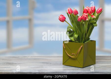 Gros plan d'un beau bouquet de tulipes rouges dans une boîte cadeau verte décorative sur une table grise rustique sur un fond de fenêtres flou. Concept de carte. Banque D'Images