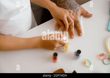 La petite fille applique du vernis jaune sur les ongles de papa à la table blanche dans la pièce Banque D'Images