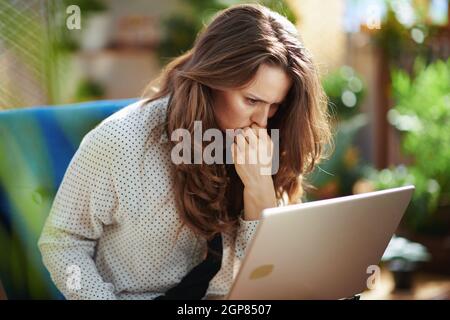 Green Home. Femme de ménage élégante et stressée avec de longs cheveux ondulés à la maison moderne en journée ensoleillée avec un ordinateur portable tout en étant assis dans un fauteuil bleu. Banque D'Images