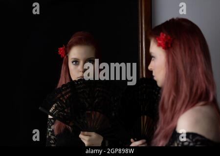 Belle jeune femme à tête rouge vêtue de vêtements flamenco et fan se regarde dans le miroir. Horizontalement. Banque D'Images