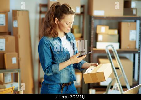 Entreprise de livraison. Femme moderne heureuse en Jean avec des colis utilisant des applications de smartphone au bureau. Banque D'Images