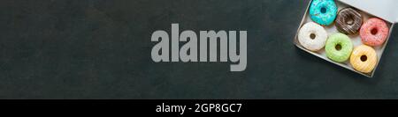 Vue du dessus de la boîte en papier avec beignets colorés et glacés sur fond texturé foncé. Longue bannière horizontale avec boîte ouverte de beignets colorés frais sur le bac Banque D'Images