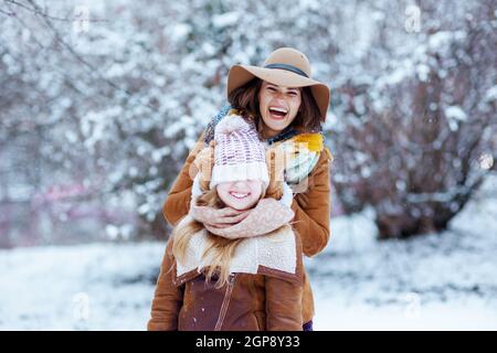 bonne mère moderne et fille en chapeaux et manteaux en peau de mouton avec des moufles jouant dehors dans le parc de la ville en hiver. Banque D'Images