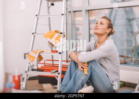 Dreamy Girl est assise sur le sol dans la plate avec un rouleau de peinture et imagine ce qu'une rénovation fraîche qu'elle fera. Réparations dans le nouvel appartement. Banque D'Images