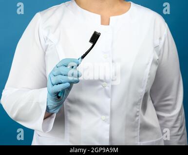 femme médecin dans un manteau médical blanc et masque se tient sur un fond bleu et tient une brosse à dents en plastique, gros plan Banque D'Images