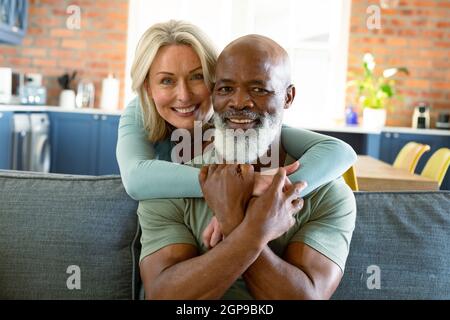 Portrait d'un heureux couple senior varié dans la salle de séjour, assis sur un canapé, embrassant et souriant Banque D'Images