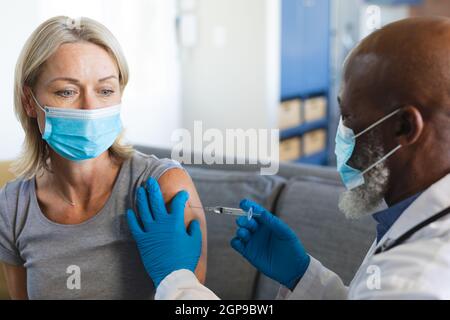 Heureuse senior diverse femme et médecin portant des masques dans la salle de séjour assis sur un canapé, vaccinant Banque D'Images
