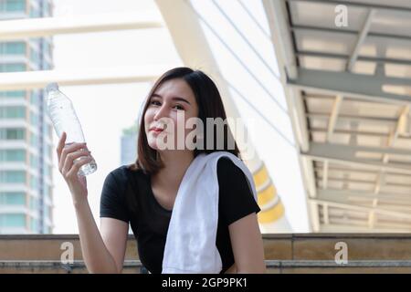 Sportive Girl avec un corps mince dans des vêtements de sport noirs et une serviette blanche sur son épaule assis sur les escaliers dans la ville moderne, tenant une bouteille d'eau minérale Banque D'Images