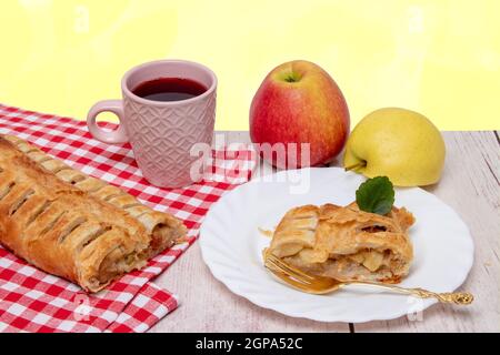 Rouleau de pommes servi. Un rouleau de strudel aux pommes frais maison avec une tasse de thé, une tranche appétissante de tarte aux pommes sur l'assiette et des pommes fraîches sur un Banque D'Images