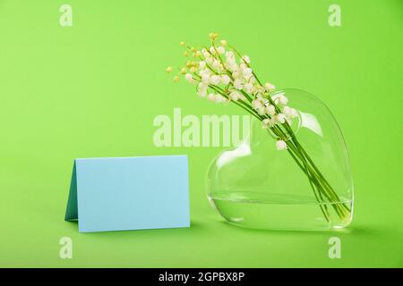 Gros plan bouquet de nénuphars de la vallée printemps frais fleurs dans un vase en verre transparent en forme de coeur et carte cadeau message sur fond vert avec co Banque D'Images