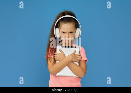 petite fille en colère dans un casque tenant une tablette numérique et crier sur fond bleu Banque D'Images