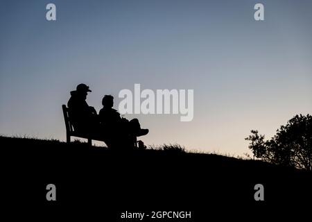 Un couple assis sur un banc de parc, silhoueté contre un ciel de crépuscule, pique-nique et profiter du coucher du soleil Banque D'Images