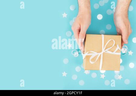 Flat Lay avec des mains adultes isolées tenant le papier artisanat boîte cadeau attaché avec ruban, étoile et cercle papier confetti ou paillettes sur fond bleu pastel Banque D'Images