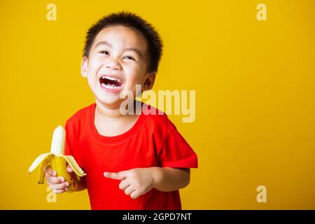 Joyeux portrait enfant ou enfant asiatique mignon petit garçon beau sourire portant un t-shirt rouge jouant tient pelé banane pour manger, studio tourné isolé sur Banque D'Images