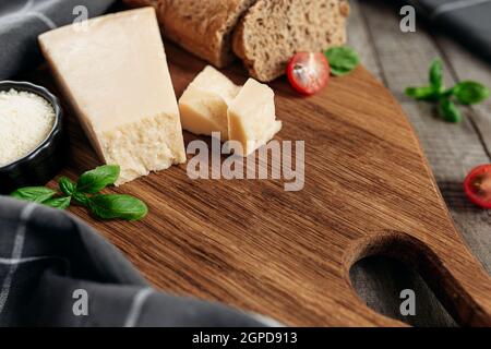 Concept de cuisine italienne. Planche à découper, morceaux de parmesan, fromage râpé dans un petit bol noir, tomate cerise, branches de feuilles de basilic, pain croustillant Banque D'Images