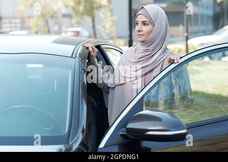 Prise de vue horizontale moyenne d'une femme musulmane confiante portant le hijab ouvrant la porte de la voiture en regardant loin Banque D'Images