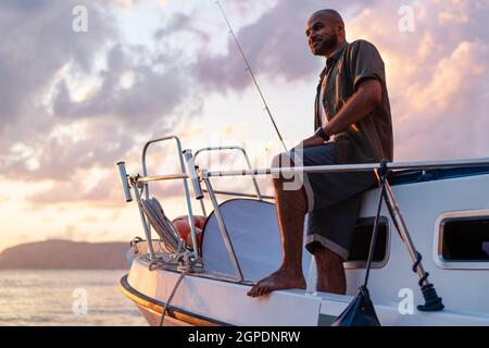 Jeune afro-américain debout avec une canne à pêche sur un voilier pêchant en pleine mer au coucher du soleil Banque D'Images