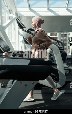 Vue latérale verticale image complète d'une femme musulmane sportive moderne portant le hijab en train de courir en salle de gym avec un tapis roulant Banque D'Images