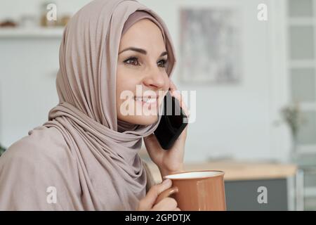 Portrait horizontal en gros plan d'une jeune femme joyeuse portant le hijab qui boit du café ou du thé et qui parle au téléphone Banque D'Images