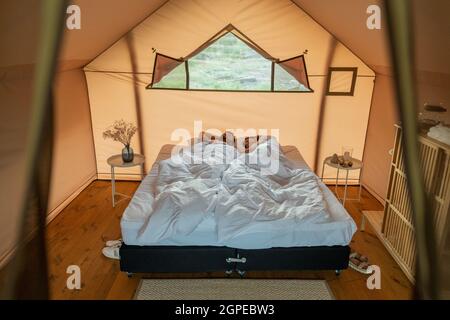 Jeune couple fatigué dormant sur un grand lit double sous des draps blancs dans une tente de glamping Banque D'Images