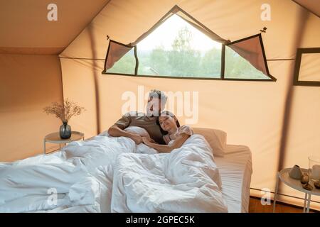 Couple affectueux assis dans un lit sous des draps blancs dans une grande chambre de tente de glamping Banque D'Images