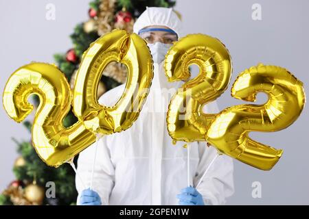 Femme en costume de protection et masque tient les numéros 2022 contre le fond de l'arbre de Noël Banque D'Images