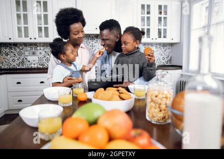 Belle famille afro-américaine de quatre personnes utilisant une tablette numérique pendant le petit déjeuner sur une cuisine lumineuse. De jeunes parents heureux regardant des dessins animés avec leurs deux filles tout en mangeant à la maison. Banque D'Images