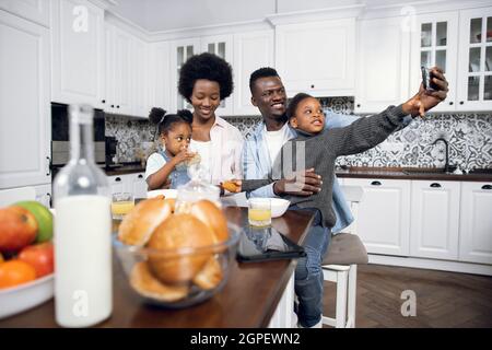 Famille afro-américaine de quatre personnes prenant le selfie sur un smartphone moderne tout en prenant le petit déjeuner sur une cuisine lumineuse. Concept des personnes, de la technologie et des modes de vie. Banque D'Images