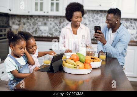 Parents afro-américains assis sur la cuisine et utilisant un smartphone tandis que leurs filles mignonnes regardant le dessin animé sur une tablette numérique. Gadgets modernes pour un style de vie. Heure du matin à la maison. Banque D'Images