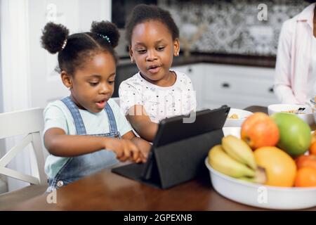 De jolies jeunes afro-américaines regardent des dessins animés sur une tablette numérique tout en étant assis ensemble dans une cuisine lumineuse. Deux sœurs utilisant des gadgets modernes pendant le petit déjeuner à la maison. Enfants et technologies concept Banque D'Images