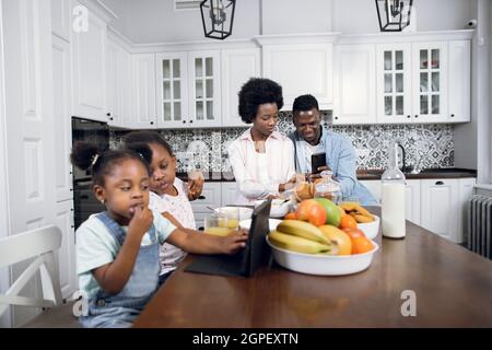 Deux sœurs africaines assises sur des chaises et regardant des dessins animés sur une tablette numérique tandis que leurs jeunes parents utilisent leur smartphone en arrière-plan. Famille avec gadgets sur la cuisine. Styles de vie modernes. Banque D'Images