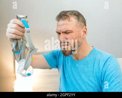 Homme heureux mature avec des problèmes respiratoires chroniques considère l'utilisation de la machine de PPC assis sur le lit dans la chambre à coucher . Santé, CPAP, ronflement concept Banque D'Images