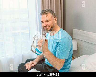 Homme heureux mature avec des problèmes respiratoires chroniques considère l'utilisation de la machine de PPC assis sur le lit dans la chambre à coucher . Santé, CPAP, ronflement concept Banque D'Images