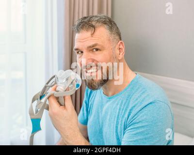 Homme heureux mature avec des problèmes respiratoires chroniques considère l'utilisation de la machine de PPC assis sur le lit dans la chambre à coucher . Santé, CPAP, ronflement concept Banque D'Images