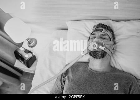 Portrait monochrome de l'homme endormi souffrant de problèmes respiratoires chroniques envisage d'utiliser la machine CPAP dans le lit. Soins de santé, thérapie obstructive d'apnée du sommeil Banque D'Images