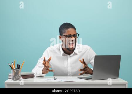Homme noir frustré assis au bureau avec un ordinateur portable et criant sur fond bleu, espace de copie Banque D'Images