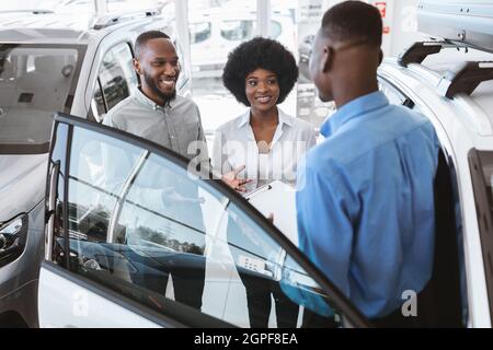 Jeune couple noir qui choisit une nouvelle voiture chez un concessionnaire automobile, un vendeur les aidant à faire le choix Banque D'Images