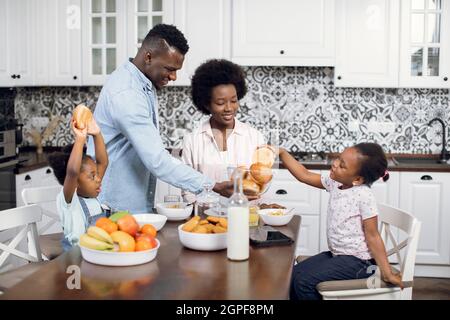 La femme et le mari afro-américains préparent un délicieux petit déjeuner pour leurs deux jolies filles. Une famille agréable et agréable qui passe le matin ensemble à la maison. Une alimentation saine. Banque D'Images