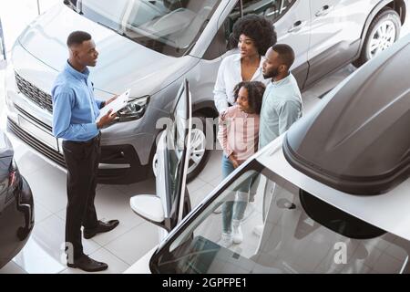 Concept commercial de vente de voitures. Vue ci-dessus du Manager parlant à la famille afro avec un enfant mignon, vendant l'automobile à la boutique de la concession Banque D'Images