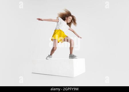 Portrait complet d'une fille souriante dans des vêtements décontractés debout sur une grande boîte isolée sur fond blanc de studio. Concept d'enfance heureuse. Banque D'Images