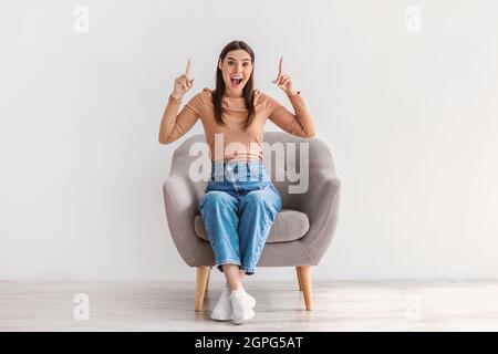 Femme millénaire surjoyeuse assise dans un fauteuil, pointant les doigts vers le haut, criant dans l'excitation, offrant de l'espace pour votre annonce Banque D'Images