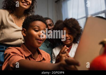 un garçon afro-américain qui fait des gestes lors d'un appel vidéo sur un ordinateur portable flou près d'une famille heureuse Banque D'Images