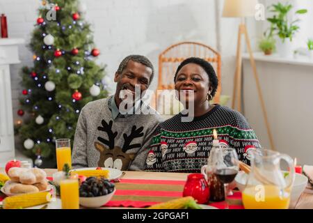 joyeux couple afro-américain près de la table servi avec un dîner de fête et un arbre de noël sur fond flou Banque D'Images