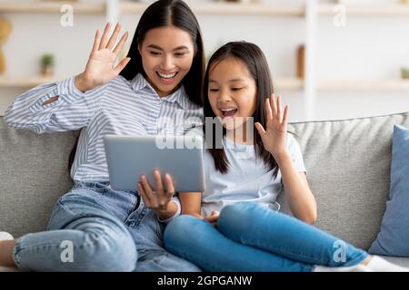 Bonne mère asiatique et fille ayant un appel vidéo sur une tablette numérique, assis sur un canapé et agitant à l'écran de pads Banque D'Images